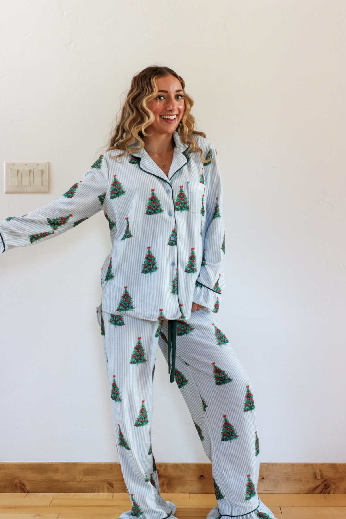 Woman wearing Christmas tree-patterned pajamas standing against a white wall.