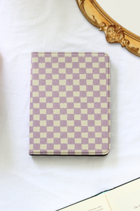A nook case with a lavender and cream checkerboard pattern, photographed on a white fabric surface with part of an ornate gold mirror frame and an open book visible around it.