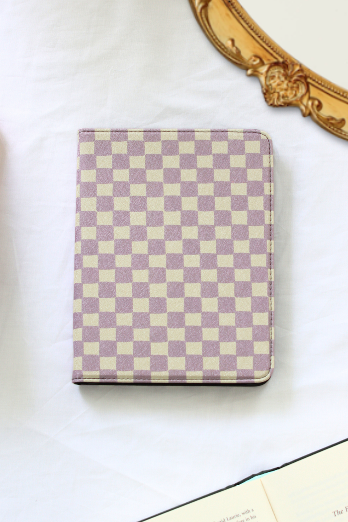 A nook case with a lavender and cream checkerboard pattern, photographed on a white fabric surface with part of an ornate gold mirror frame and an open book visible around it.