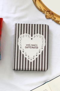 A kindle case with a brown and white striped cover, featuring a white lace-patterned heart graphic in the center with the text 'well read & CAFFEINATED' in dark red. The kindle case is photographed on a white fabric background with decorative props, including a gold mirror frame and an open book.