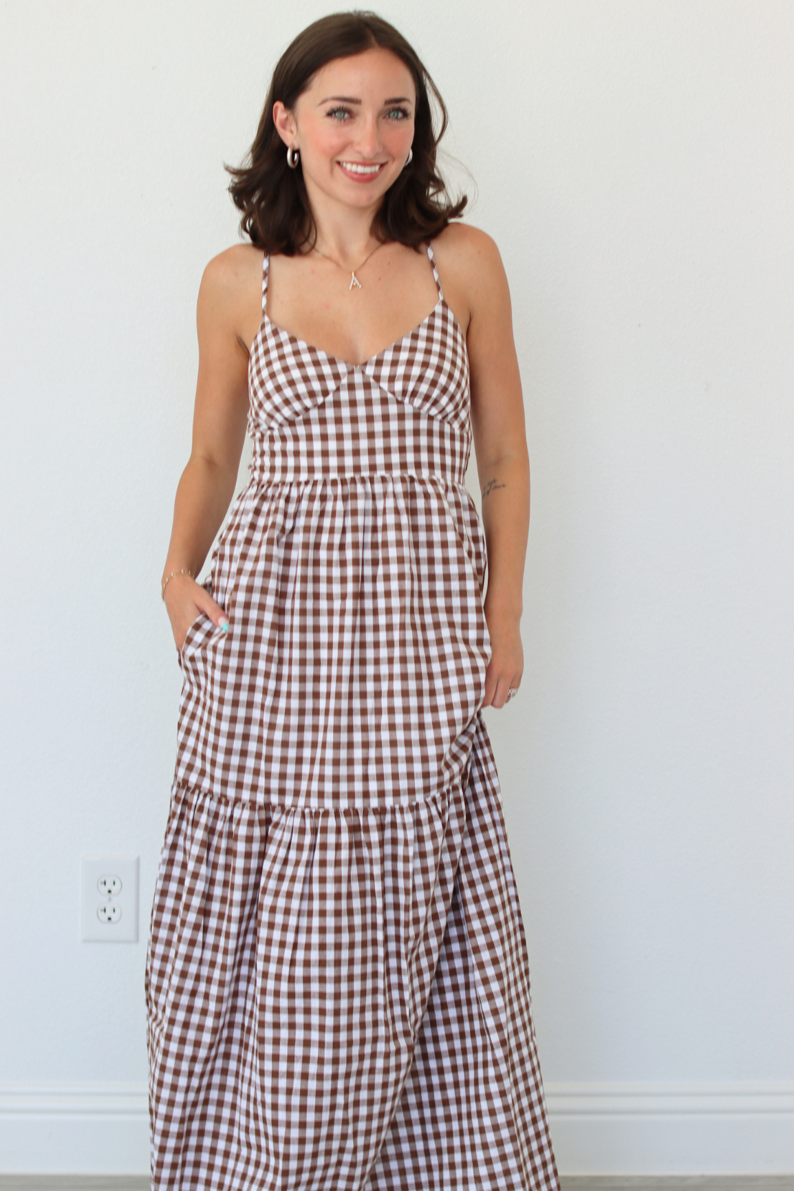 A woman modeling a brown and white gingham tiered maxi dress against a plain white wall.