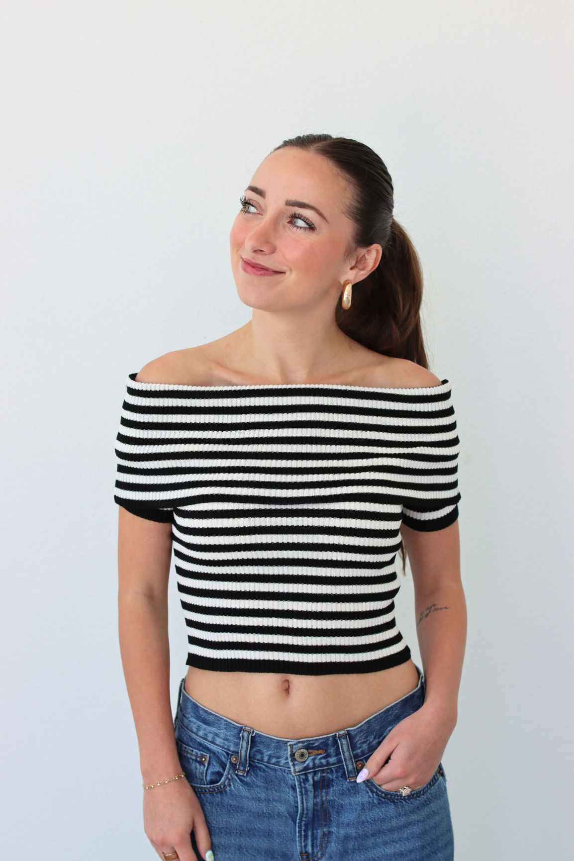 Woman wearing a black and white striped off-shoulder top and blue jeans against a plain background