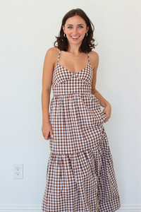 A woman modeling a brown and white gingham tiered maxi dress against a plain white wall.