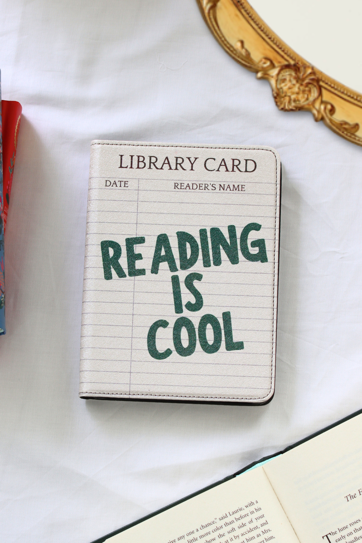 A kindle case with lined paper print and the phrase 'READING IS COOL' in dark green lettering, styled on a white fabric surface with partial books and a gold mirror frame visible.