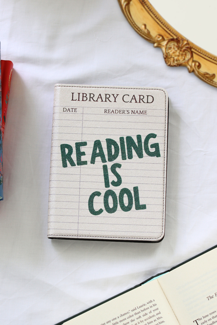 A kindle case with lined paper print and the phrase 'READING IS COOL' in dark green lettering, styled on a white fabric surface with partial books and a gold mirror frame visible.