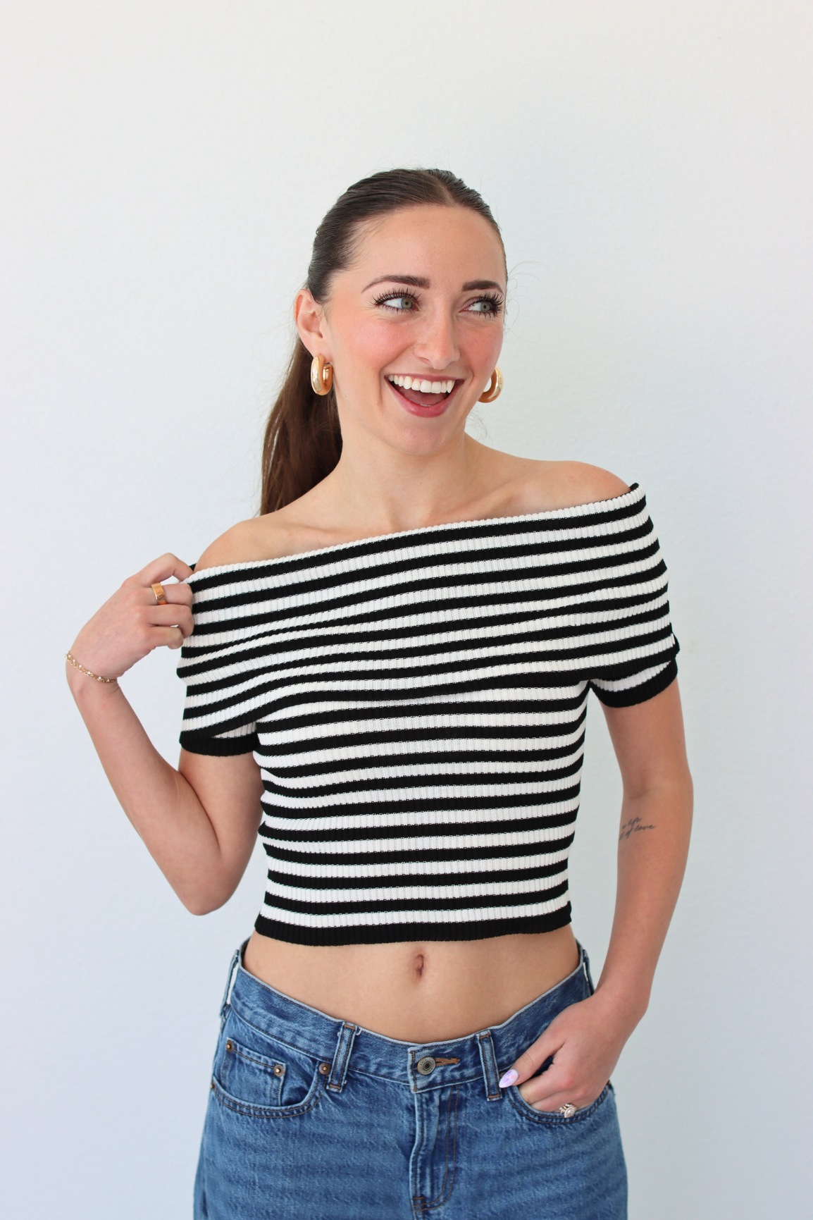 Woman wearing a black and white striped off-shoulder top and blue jeans against a plain background
