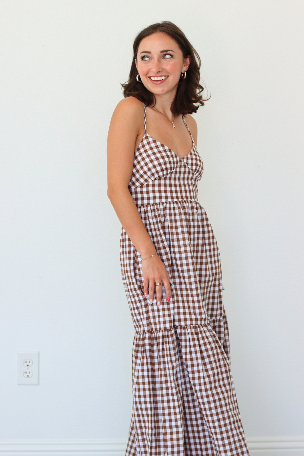 A woman modeling a brown and white gingham tiered maxi dress against a plain white wall.