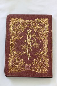 A kindle case featuring an embossed gold design of a dagger wrapped in a snake, surrounded by floral borders, with 'LND' embossed at the bottom.