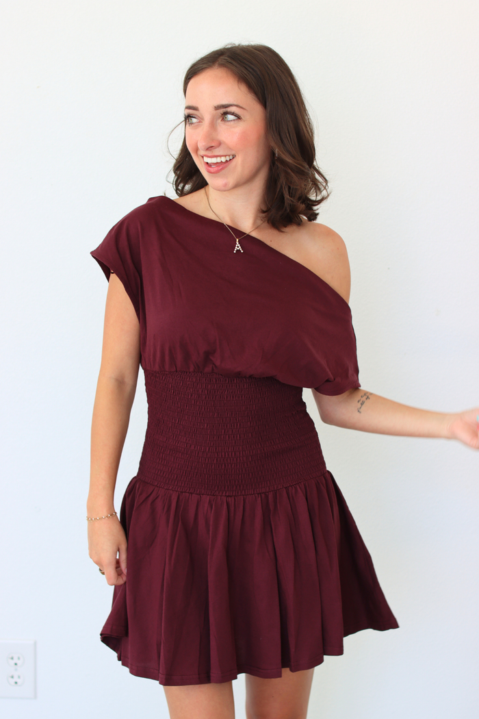 A woman modeling an off-the-shoulder mini dress in burgundy. The dress features an asymmetrical draped neckline, a smocked waist, and a flared skirt, shown against a plain white wall.