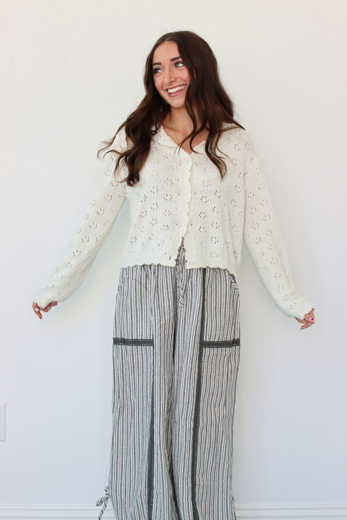 A woman modeling a cream eyelet knit cardigan over a white top and gray-and-white vertically striped wide-leg pants, standing against a plain white wall.