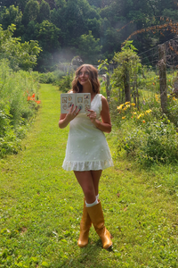 A woman stands on a grassy path in a lush garden, wearing a white sleeveless dress and mustard yellow rain boots while holding a small printed card.