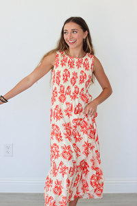 A woman modeling a sleeveless, tiered maxi dress with a red abstract floral print on a cream background, standing against a plain white wall.
