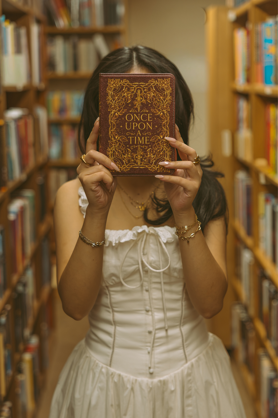 A woman in a white corset dress holds a vintage-style book titled 'Once Upon a Time' in front of her face, standing in a book-filled library aisle.
