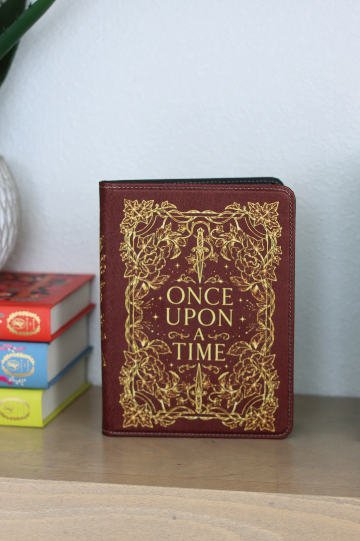 A kindle case standing upright on a wooden shelf, featuring a burgundy cover with ornate gold filigree and the words 'ONCE UPON A TIME' in gold lettering. A stack of colorful books sits to the left, and a light-colored wall is behind.
