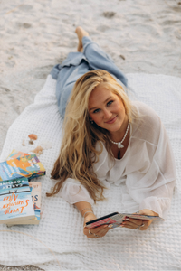 A woman with long blonde hair lies on a white blanket on a sandy beach, wearing a white blouse and blue jeans, holding a kindle case. Books are stacked on the blanket next to her.