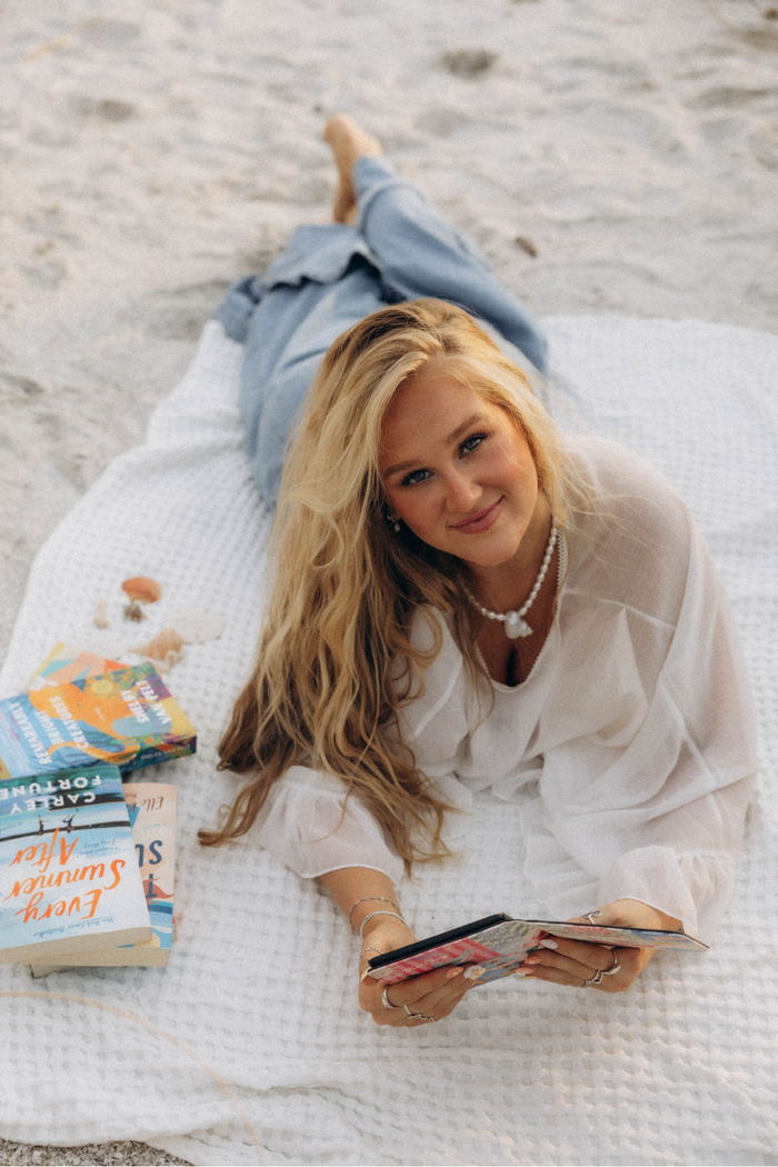 A woman with long blonde hair lies on a white blanket on a sandy beach, wearing a white blouse and blue jeans, holding a kindle case. Books are stacked on the blanket next to her.