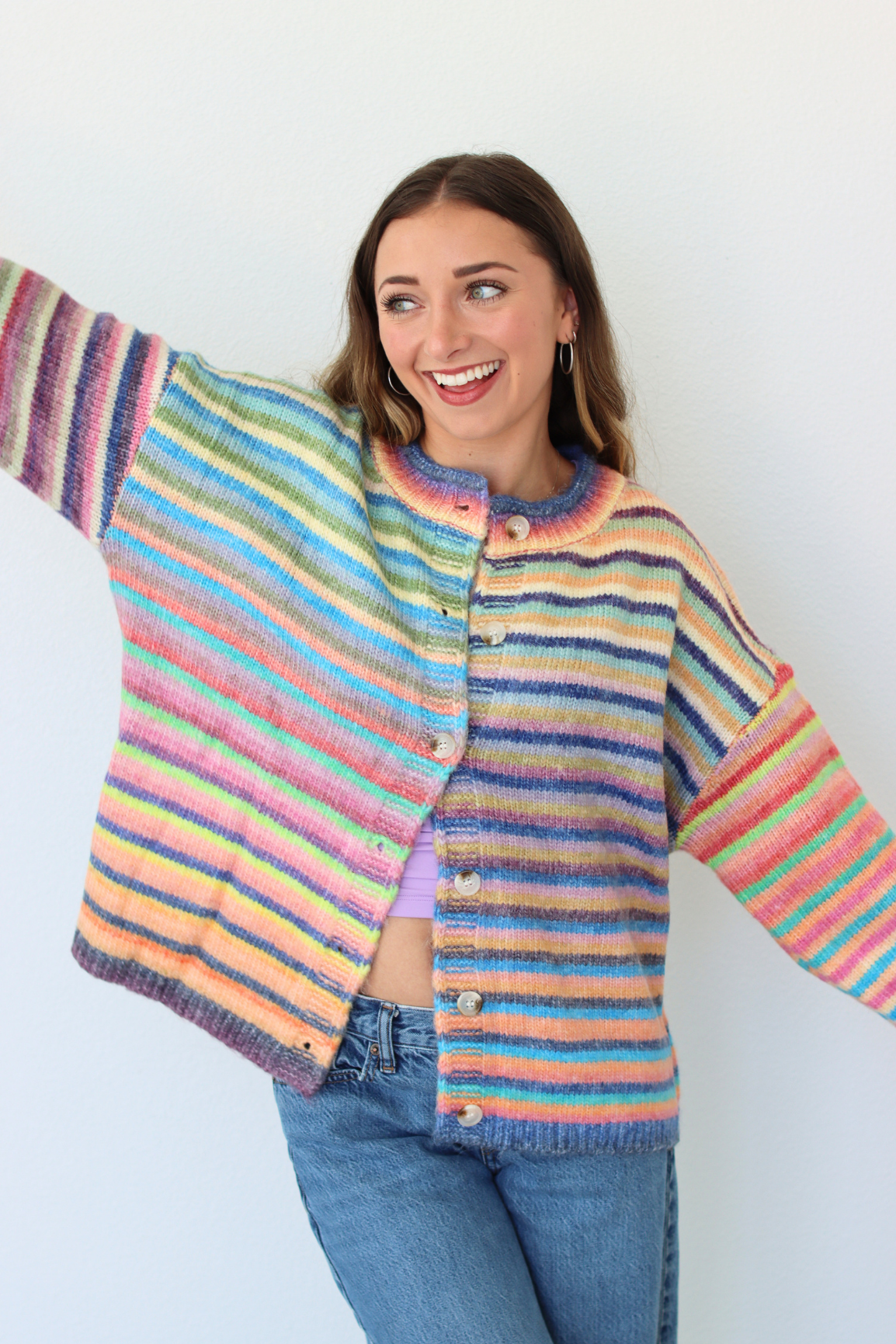 A woman wearing a colorful striped knit cardigan sweater with blue jeans, posing with one arm outstretched against a plain white background.