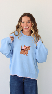 Woman wearing a light blue sweater with a cat embroidery design against a white background 