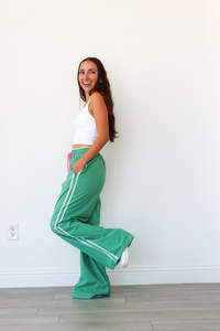 Woman wearing a white top and green pants with white stripes against a white wall.