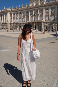 A woman stands in a large paved plaza in front of a grand classical building, wearing a white sleeveless midi dress with small black polka dots, a black cap, and carrying a white shoulder bag.
