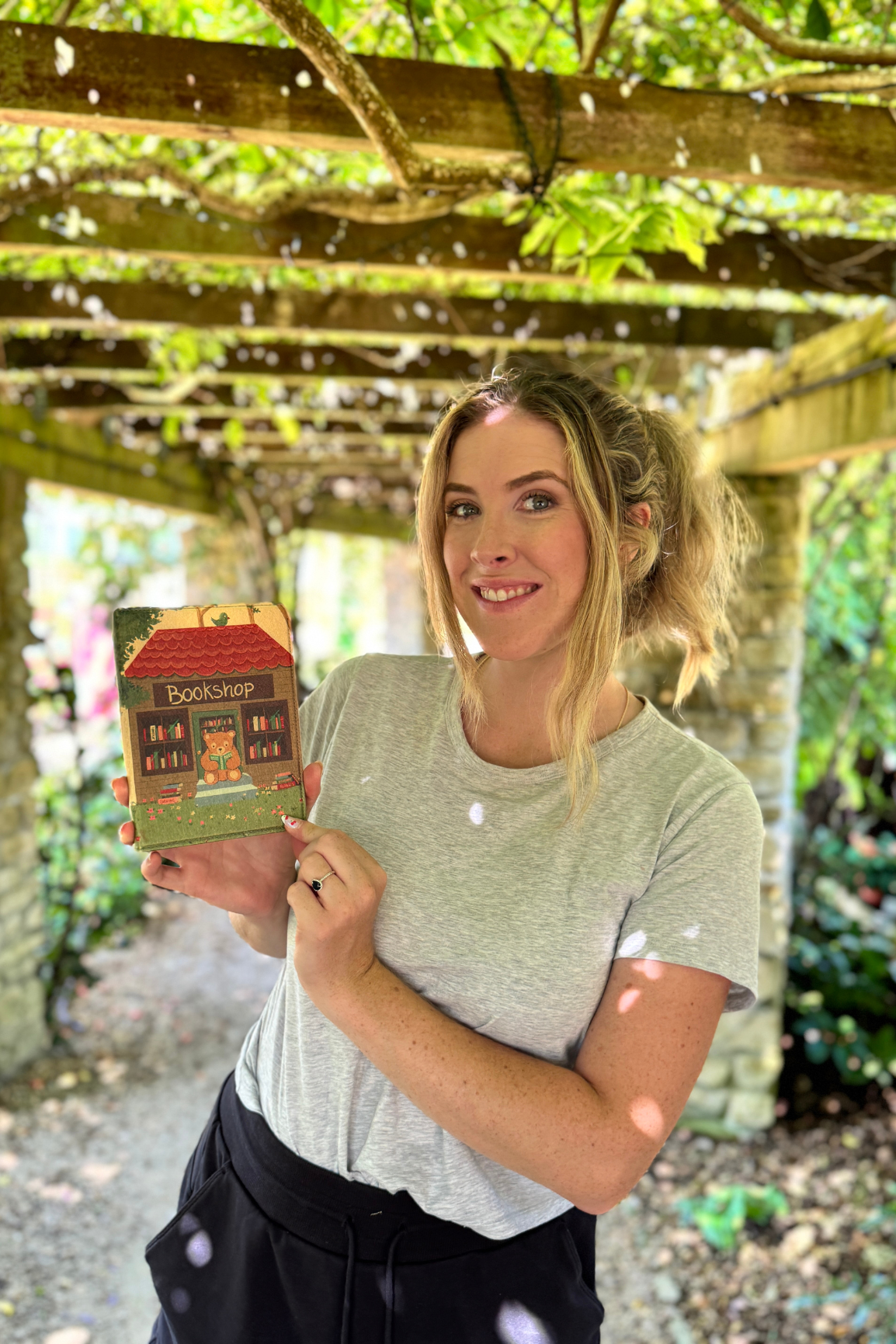 A woman smiling and holding a kindle case under a wooden pergola with green foliage overhead.