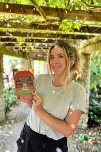 A woman smiling and holding a kindle case under a wooden pergola with green foliage overhead.