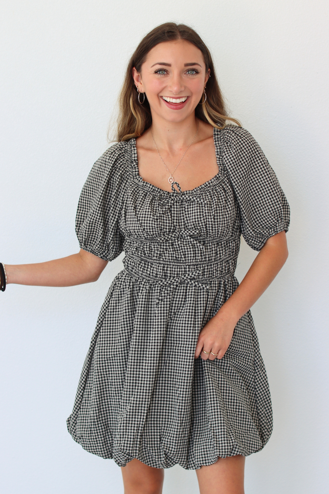 A woman modeling a black and white gingham mini dress with puff sleeves, a square neckline, and a smocked waist, set against a plain white background.