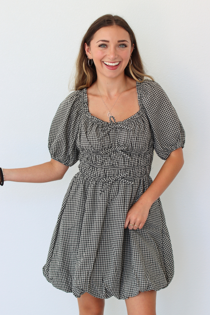 A woman modeling a black and white gingham mini dress with puff sleeves, a square neckline, and a smocked waist, set against a plain white background.