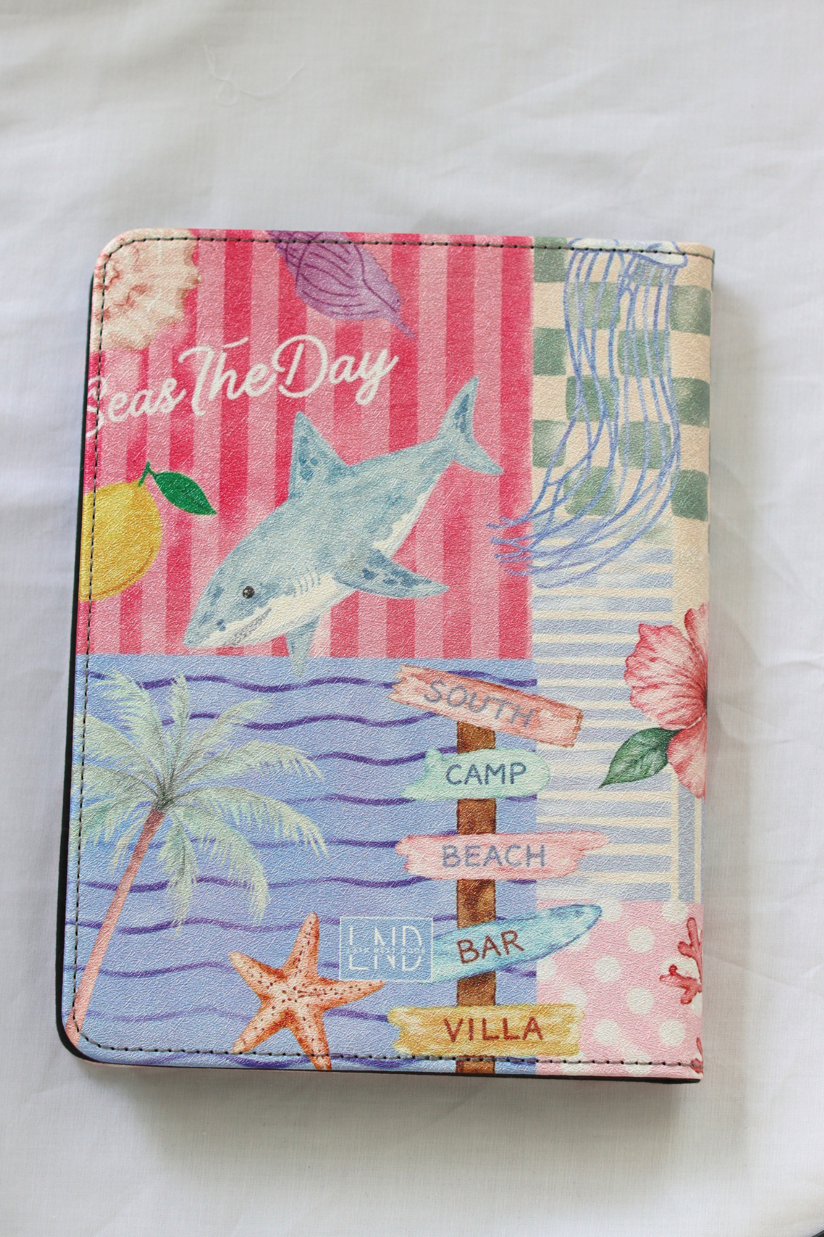 A kindle case featuring a colorful beach-themed collage with a shark, palm tree, starfish, flowers, striped background, and directional signs reading South Camp Beach and Land Bar Villa.
