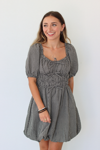 A woman modeling a short black and white gingham dress with puff sleeves, a square neckline, and a gathered smocked waist, standing against a plain white wall.