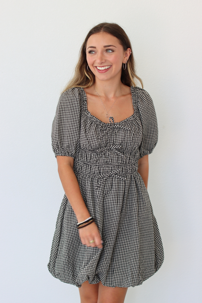A woman modeling a short black and white gingham dress with puff sleeves, a square neckline, and a gathered smocked waist, standing against a plain white wall.