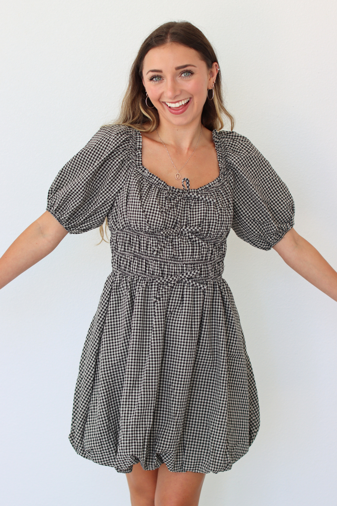 A woman modeling a black and white gingham mini dress with puff sleeves, a smocked bodice, and a gathered skirt, set against a plain white background.