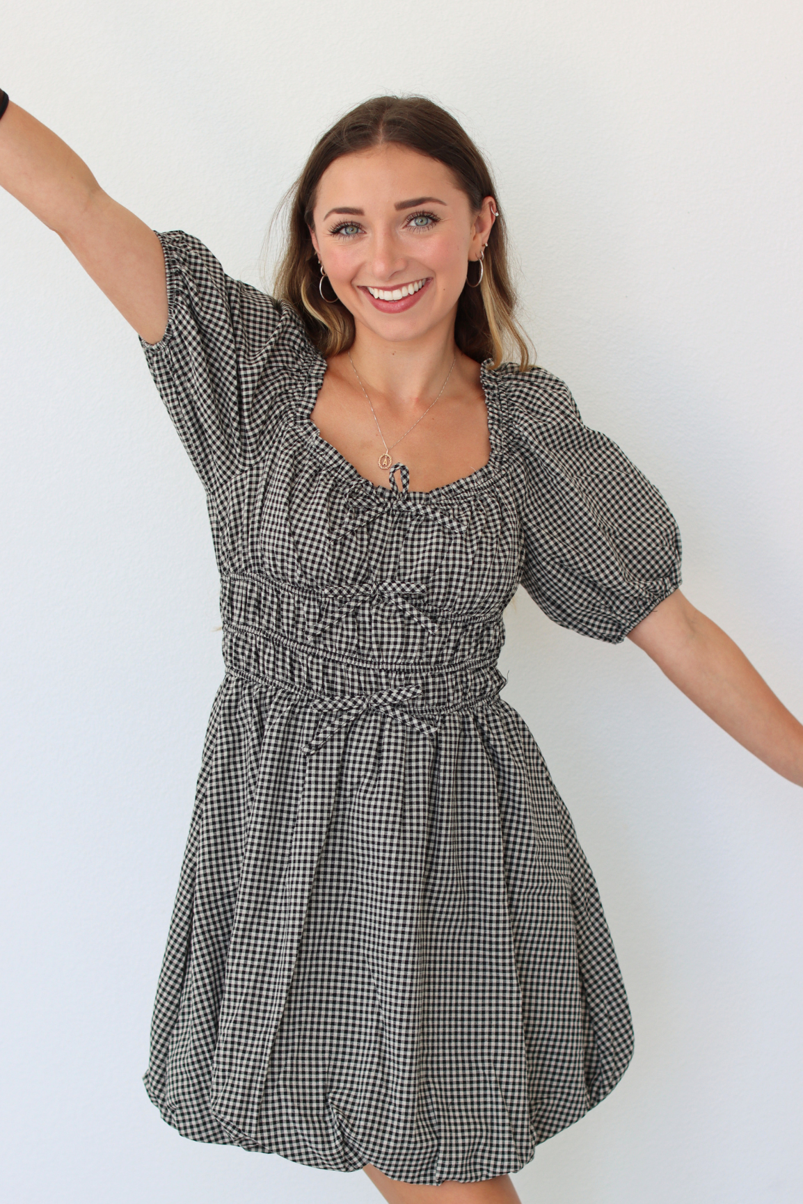 A woman modeling a black and white gingham mini dress with puff sleeves, a smocked waist, and a square neckline, posing with her arms outstretched against a plain white wall.