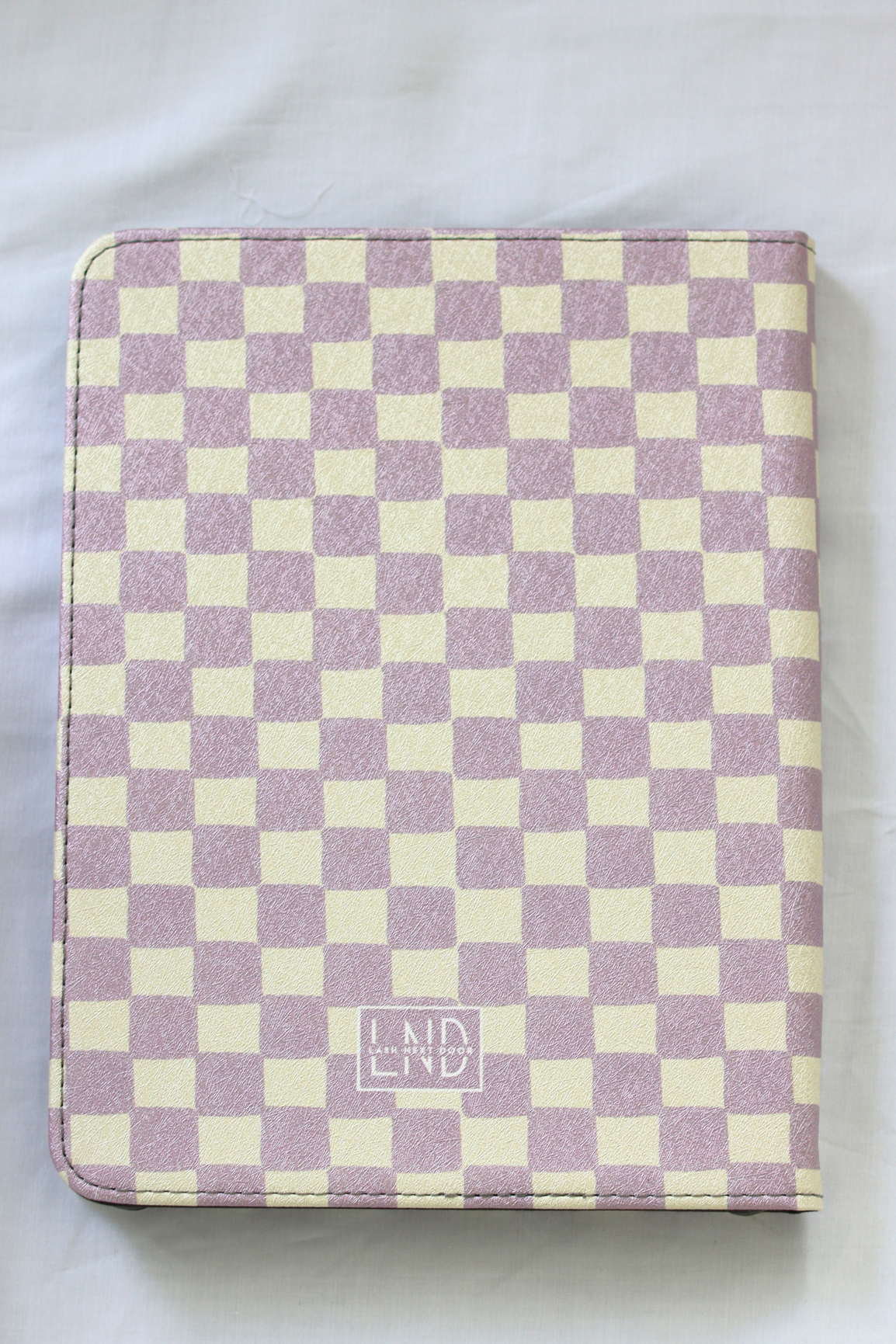 A nook case featuring a lavender and cream checkerboard pattern, with a small 'LND' logo near the bottom center, laid flat on a white fabric background.