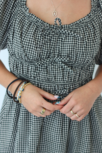 A close-up of a person wearing a black and white gingham dress with a smocked bodice and tie-front detail. The person is also wearing multiple bracelets and rings.