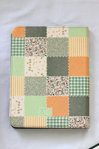 A nook case featuring a patchwork quilted design with various fabric patterns in green, orange, beige, and gray, laid flat on a white background.