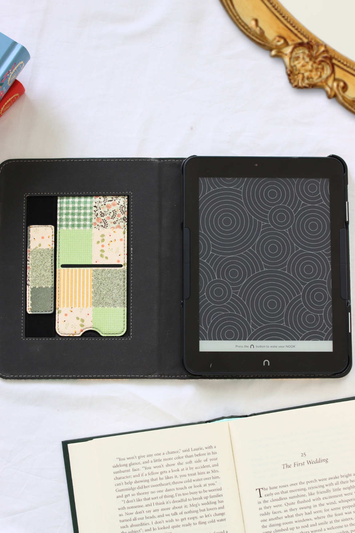 A nook case shown open on a white marble-like surface, with a patterned card holder insert on the left and a black tablet screen on the right. An open book is visible at the bottom, and part of a gold mirror frame and colorful objects appear at the edges.