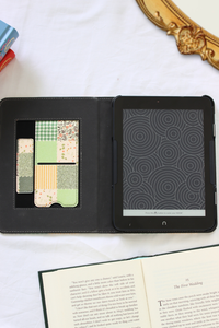 A nook case shown open on a white marble-like surface, with a patterned card holder insert on the left and a black tablet screen on the right. An open book is visible at the bottom, and part of a gold mirror frame and colorful objects appear at the edges.
