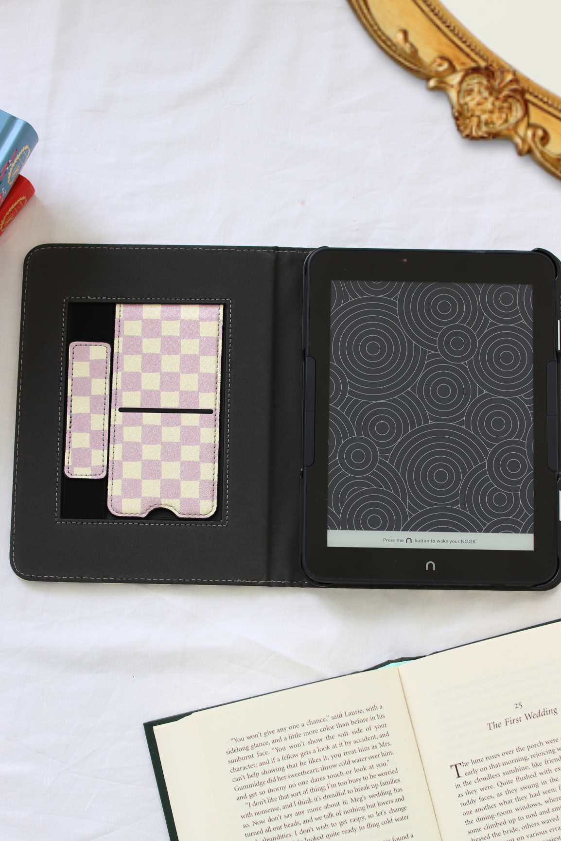 A nook case shown open on a white fabric surface, with a matching strap and kickstand. A tablet is inserted, and an open book is partly visible at the bottom.