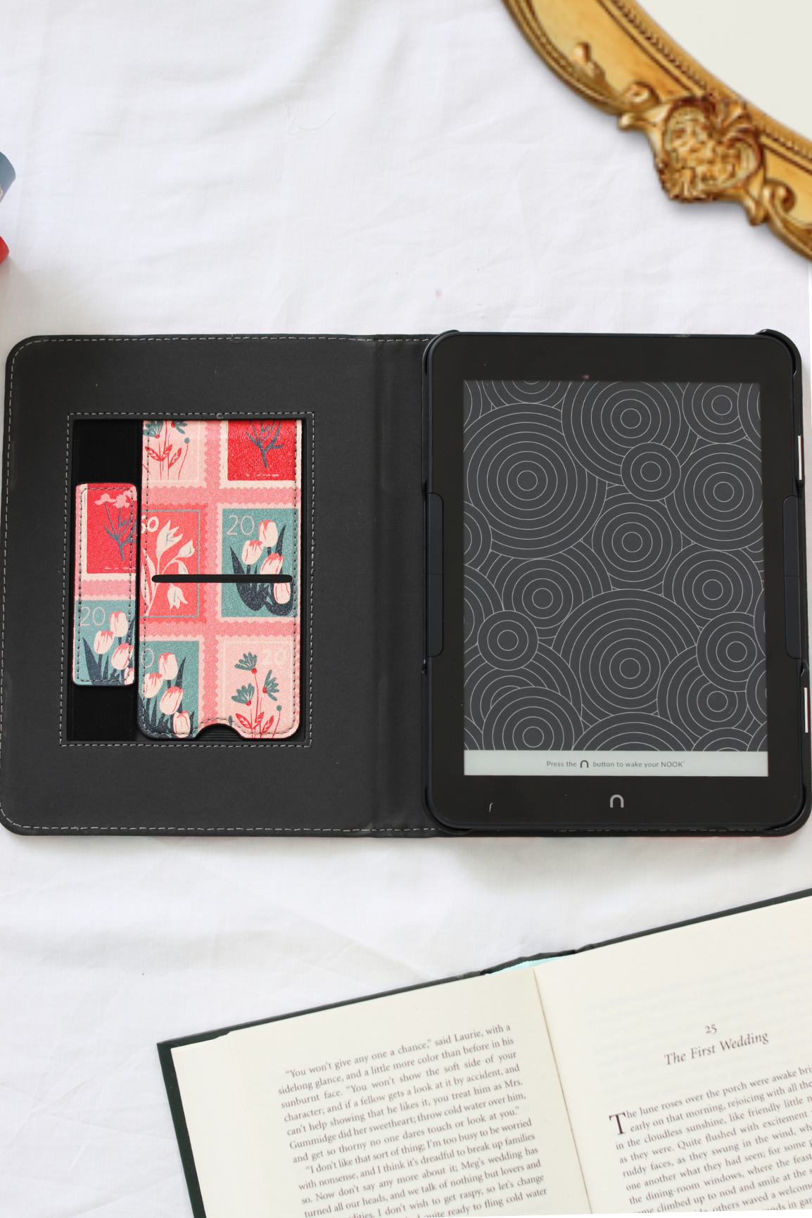 A nook case shown open on a white fabric surface, with a tablet inserted on the right side and card slots with floral-patterned cards on the left. An open book is visible at the bottom, and part of a gold mirror frame appears at the top right.