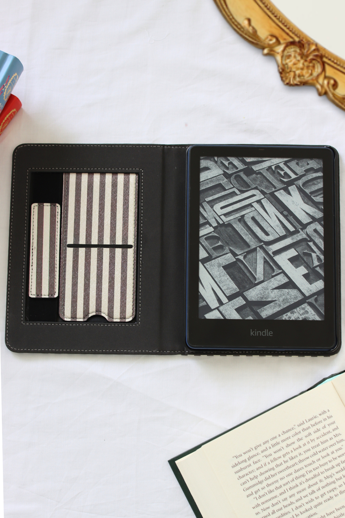 A Kindle e-reader folio case shown open on a white fabric surface, with card slots on the left and an e-reader screen on the right. Decorative props including an open book, a gold mirror edge, and colored pencils are partially visible around it.
