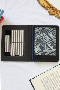 A Kindle e-reader folio case shown open on a white fabric surface, with card slots on the left and an e-reader screen on the right. Decorative props including an open book, a gold mirror edge, and colored pencils are partially visible around it.