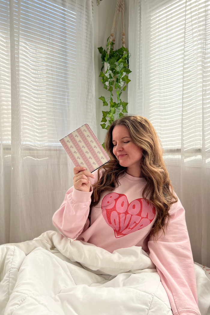 Woman in pink sweatshirt with heart design holding a pink kindle in a room with white curtains and green plant.