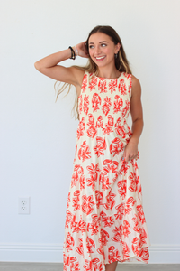 A woman modeling a sleeveless, tiered maxi dress with a red abstract floral print on a cream background, standing against a plain white wall.