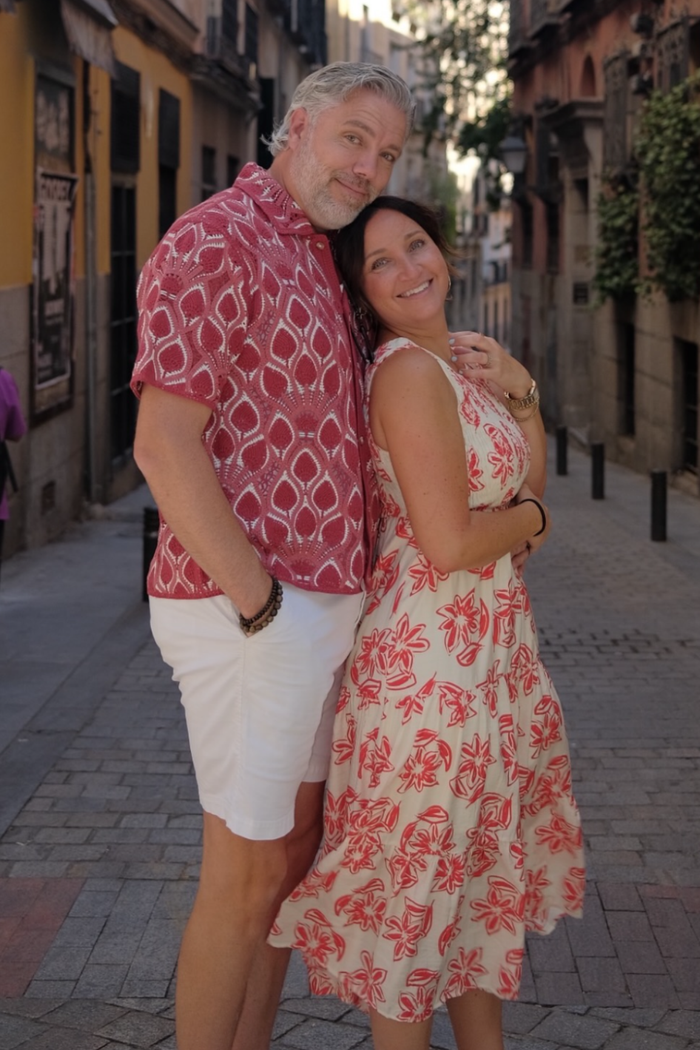 A couple posing together on a cobblestone street. The man wears a red and white patterned short-sleeve button-up shirt and white shorts, while the woman wears a sleeveless cream dress with a red floral pattern.