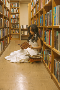 A woman in a white dress sits on the floor of a library aisle, reading a book surrounded by tall wooden bookshelves filled with books.