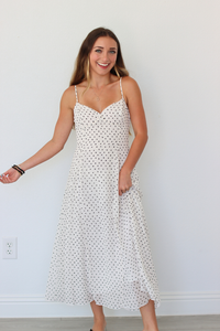 A woman modeling a white spaghetti-strap midi dress with a small black polka dot pattern, standing against a plain white wall.