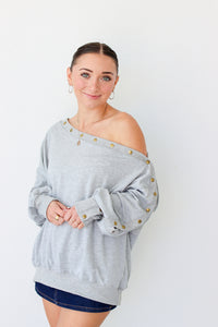 Woman wearing a light gray off the shoulder sweater with buttons along the trim 