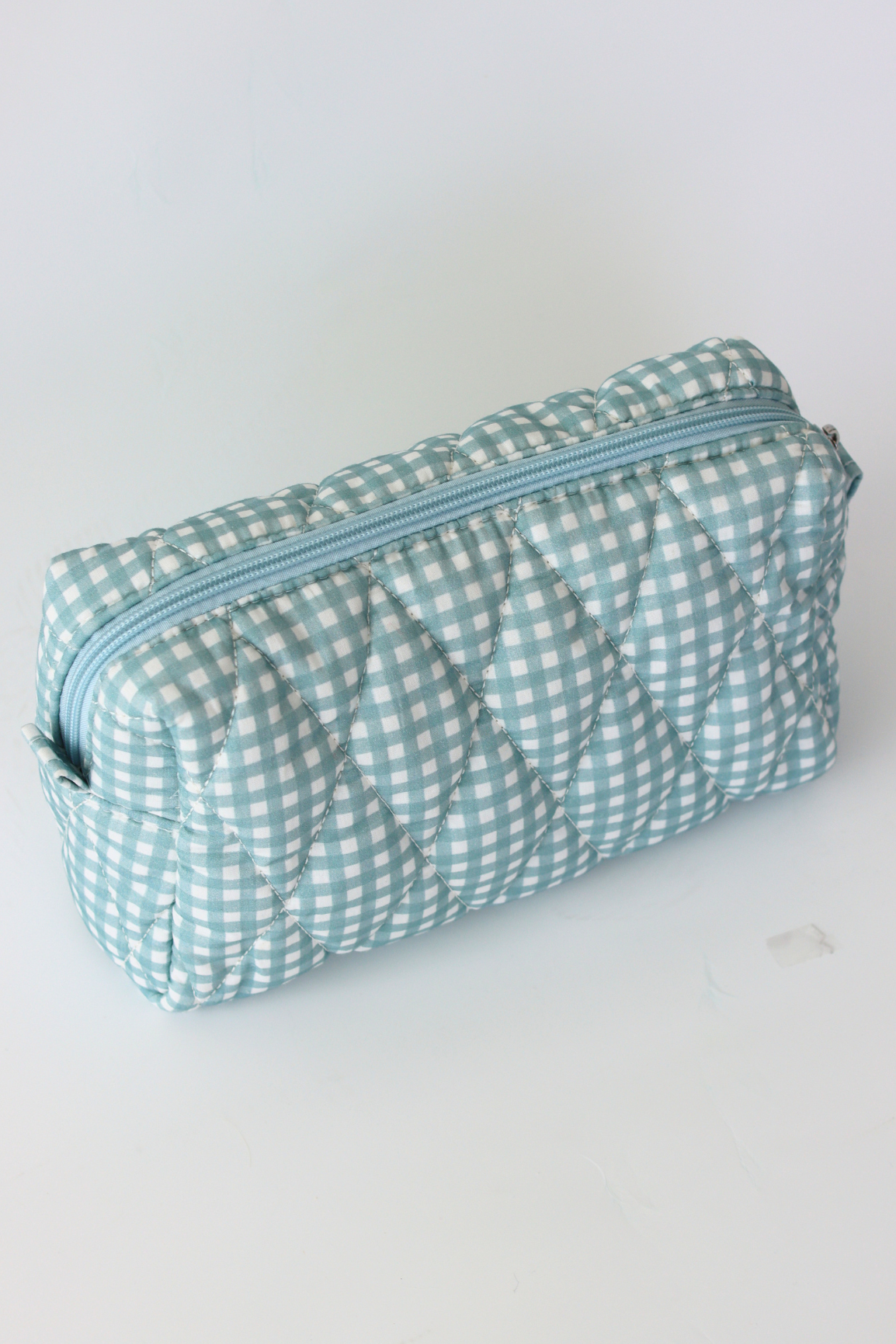 A quilted cosmetic pouch with a light blue and white gingham pattern, featuring a diagonal zipper, shown on a plain white background.