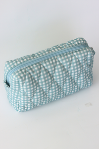 A quilted cosmetic pouch with a light blue and white gingham pattern, featuring a diagonal zipper, shown on a plain white background.